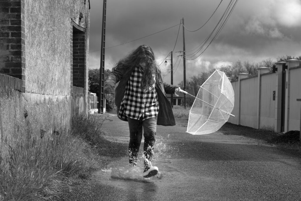 Petite fille avec un parapluie jouant dans une flaque d’eau, photo en noir et blanc au style poétique inspiré de Robert Doisneau.