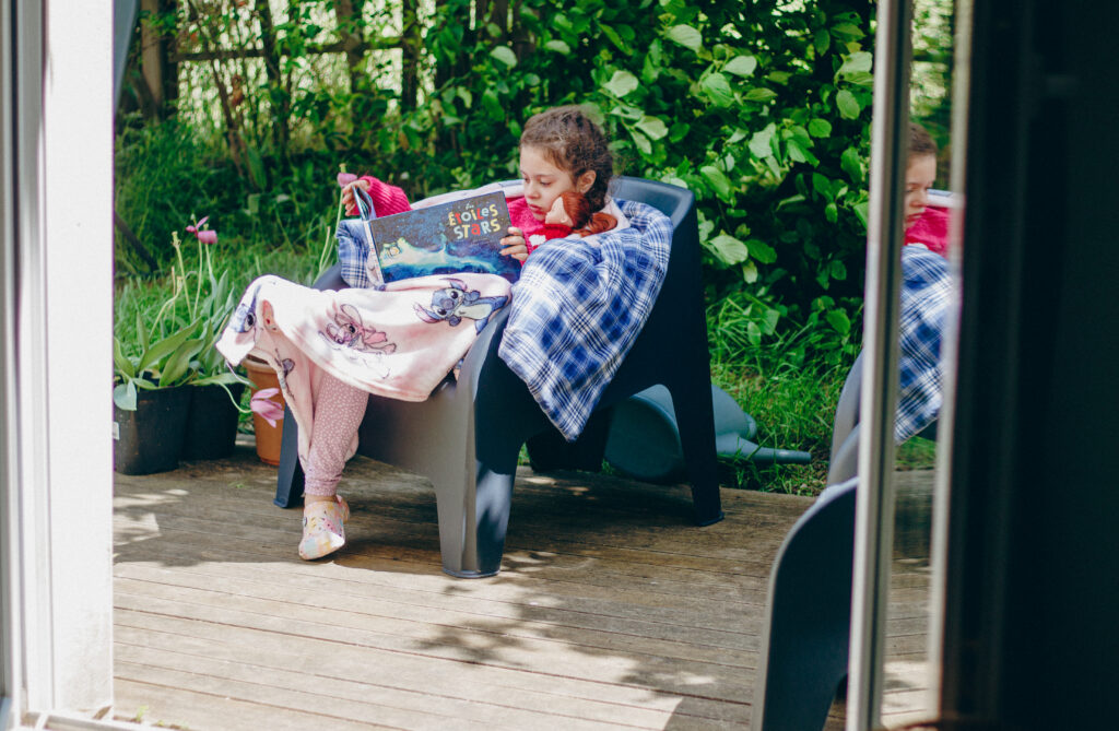 Petite fille lisant tranquillement sur une terrasse baignée de lumière, un doux glimmer de mai dans une ambiance calme et chaleureuse.